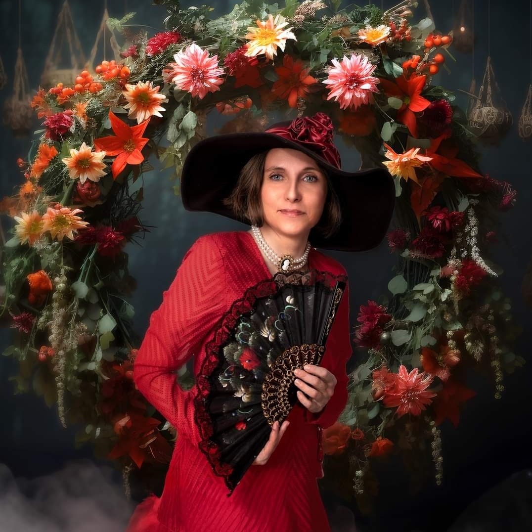 Portret kobiety w czerwonej sukience i kapeluszu z szerokim rondem, trzymającej ozdobny wachlarz na tle kwiatowego łuku