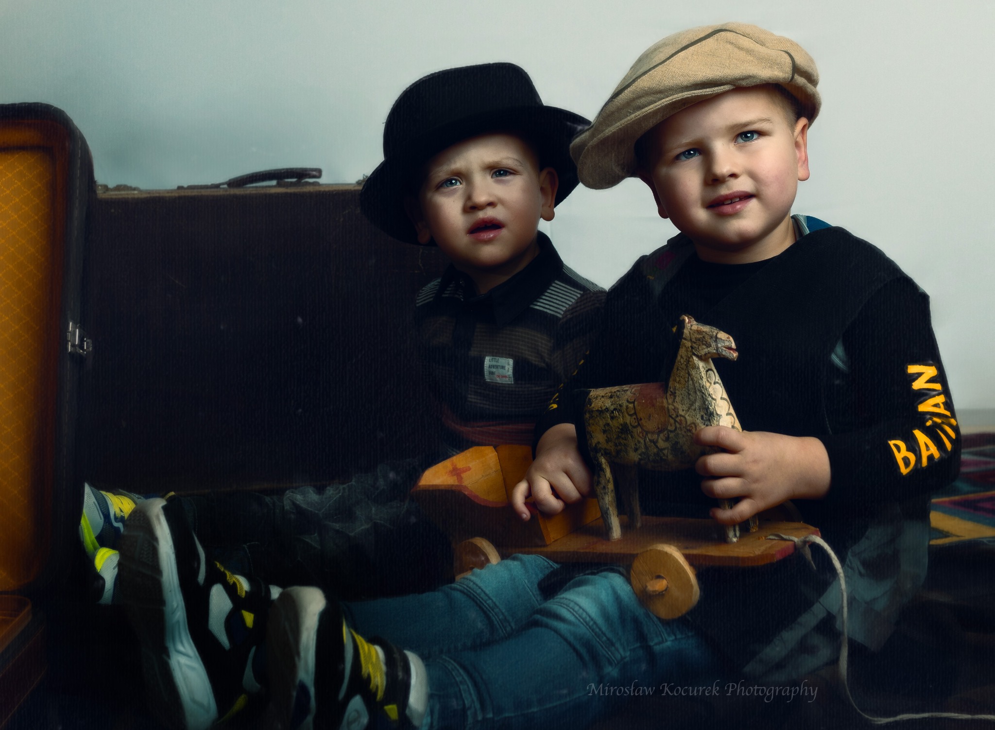 Dwóch chłopców w kapeluszach pozuje z drewnianym koniem na kółkach przed starą walizką.