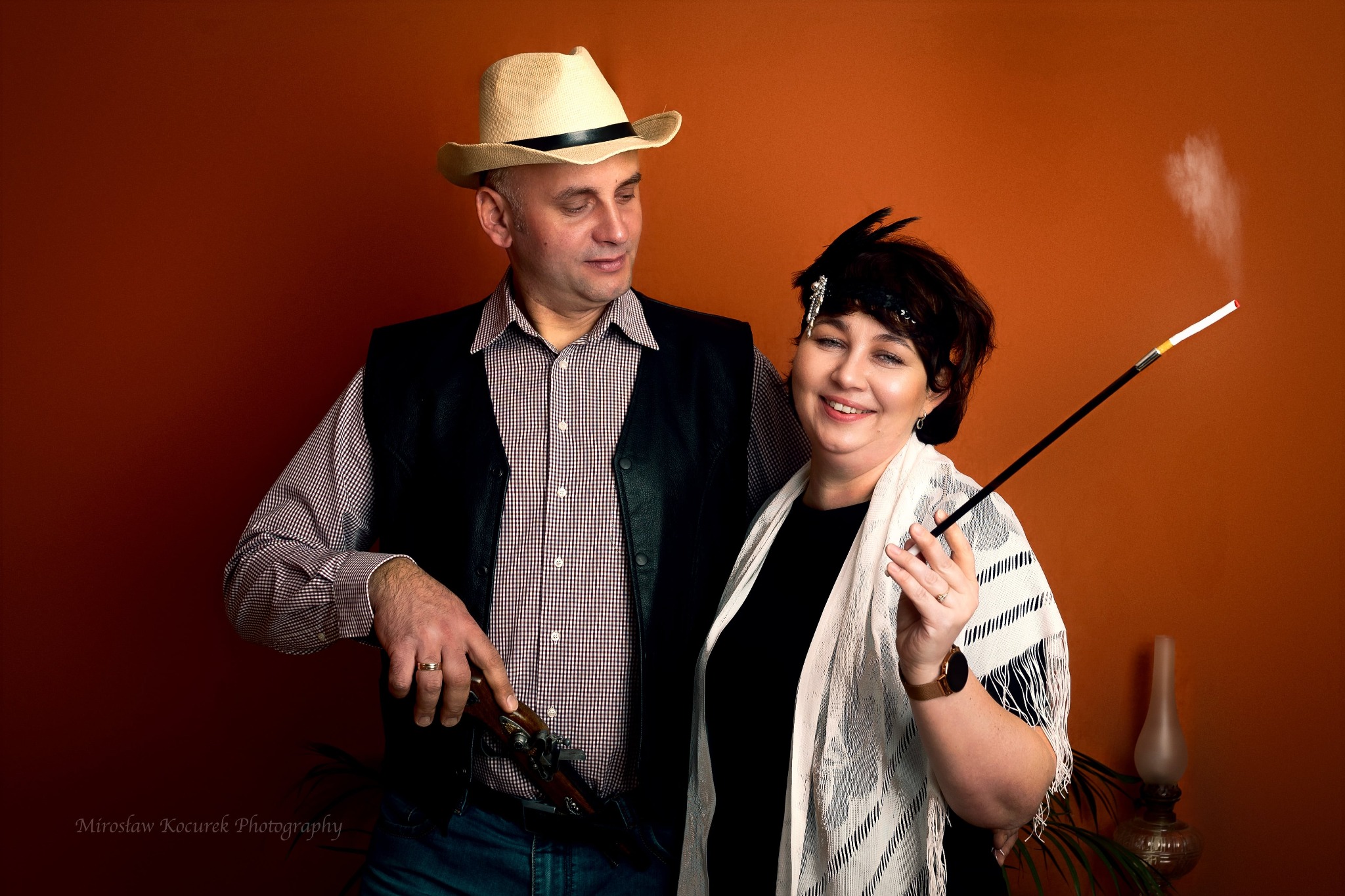 Para w stylizowanych strojach: mężczyzna w kapeluszu i kamizelce trzymający starodawną broń, kobieta z cygaretką w lufce, w opasce z piórkiem i szalu na tle pomarańczowej ściany.