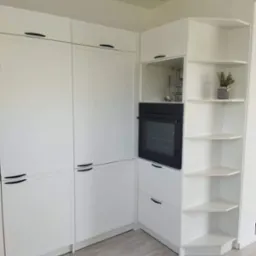 Nowoczesna, biała zabudowa kuchenna z czarnymi, minimalistycznymi uchwytami, piekarnikiem w zabudowie i narożnymi półkami z dekoracyjną rośliną w doniczce.