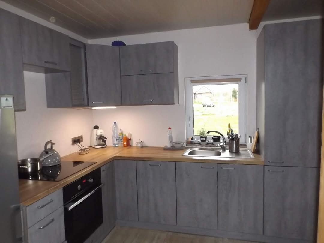 Szara kuchnia z drewnianym blatem, widok na okno z widokiem na ogród, czajnik na płycie indukcyjnej i ekspres do kawy na blacie.