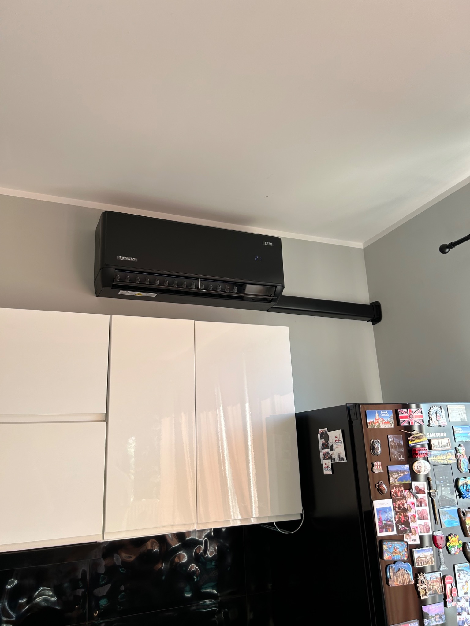 Czarny klimatyzator Rotenso zamontowany na szarej ścianie nad białymi szafkami kuchennymi i czarną lodówką ozdobioną magnesami.