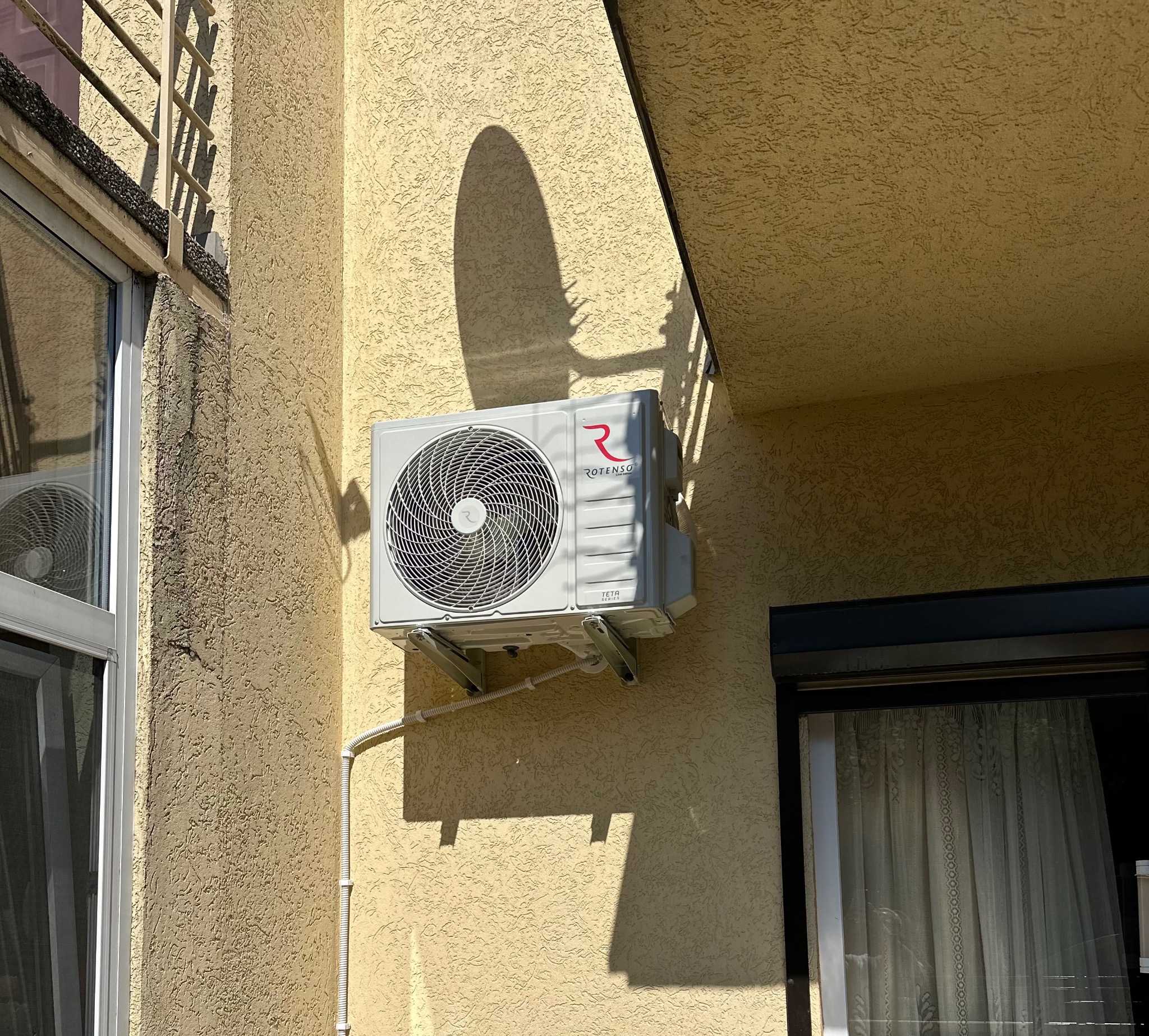 Zewnętrzna jednostka klimatyzacji Rotenso Teta Series zamontowana na elewacji domu z oknem i roletą, widoczne przewody i cień urządzenia.