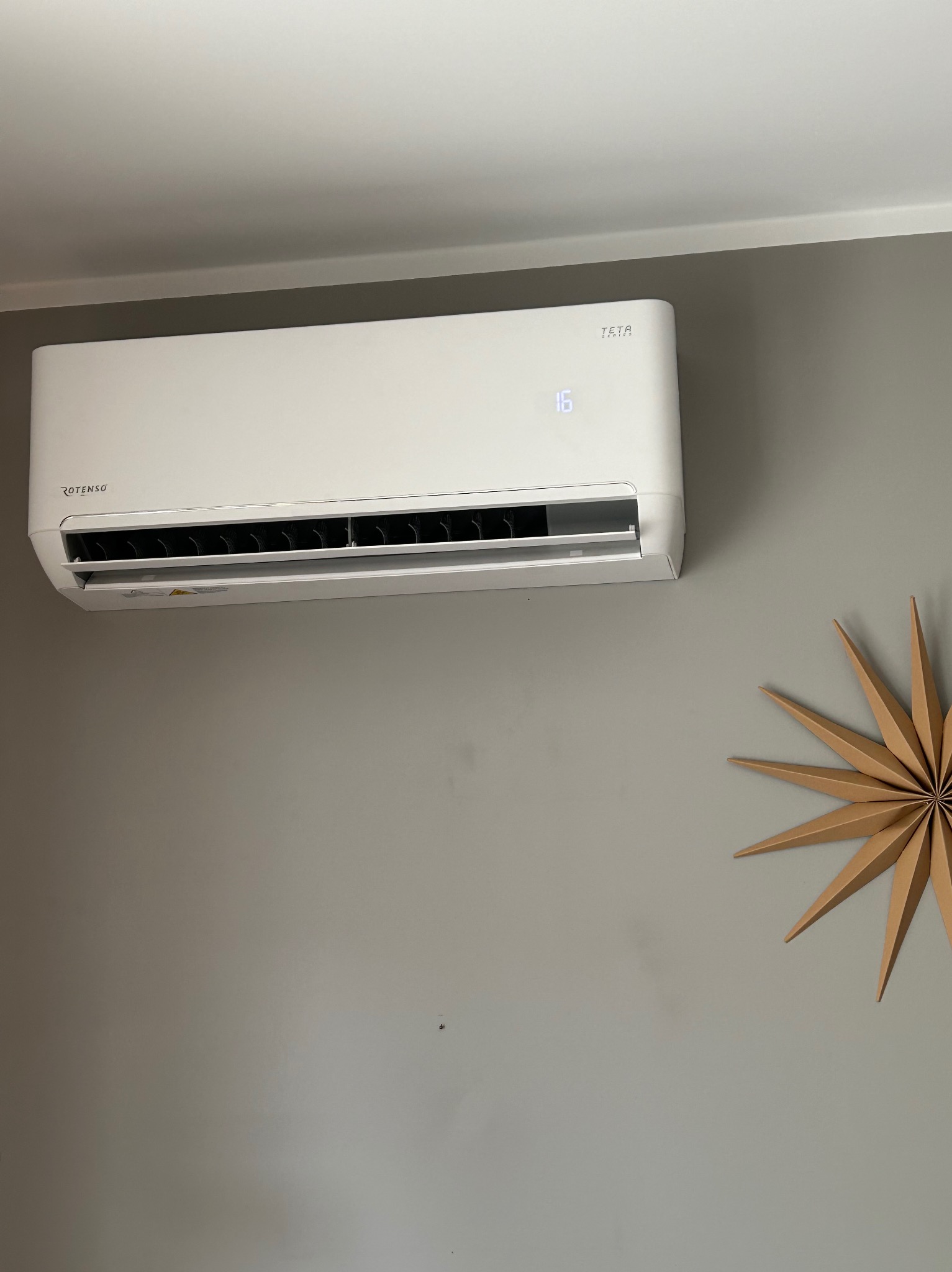 Biały klimatyzator Rotenso z wyświetlaczem temperatury 16 stopni Celsjusza, zamontowany na szarej ścianie w pomieszczeniu z dekoracyjną gwiazdą z papieru w tle.