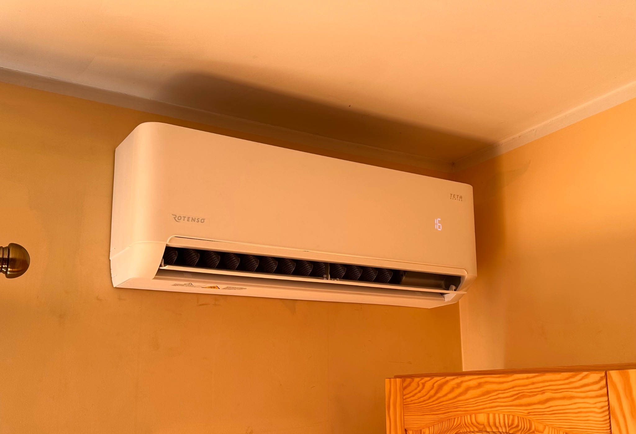 Biały klimatyzator Rotenso Teta Series zamontowany na pomarańczowej ścianie w domu, widoczny fragment drewnianego mebla.