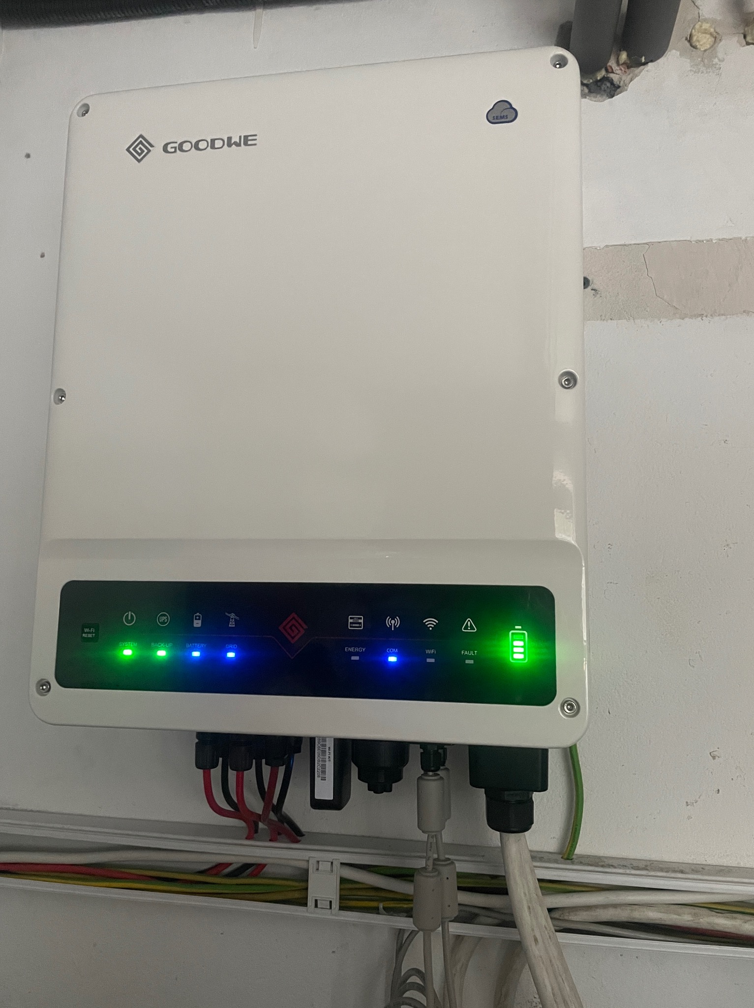 Biała obudowa inwertera Goodwe z podłączonymi przewodami, widoczne diody sygnalizacyjne: zielone dla systemu i energii, niebieskie dla baterii, WiFi i komunikacji.