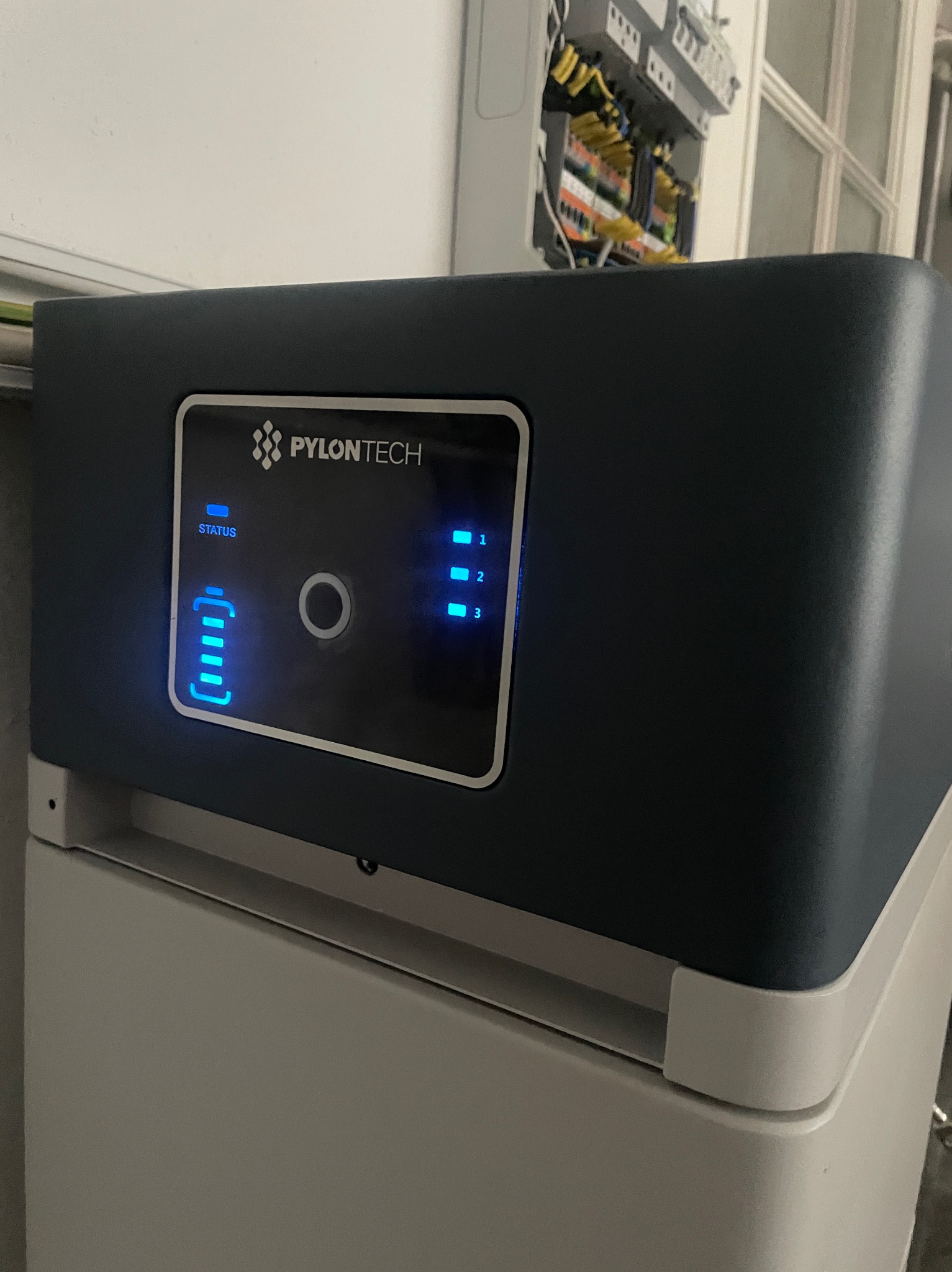 Szary magazyn energii Pylontech z niebieskimi diodami LED wskazującymi poziom naładowania i status, widoczna skrzynka elektryczna z okablowaniem w tle.