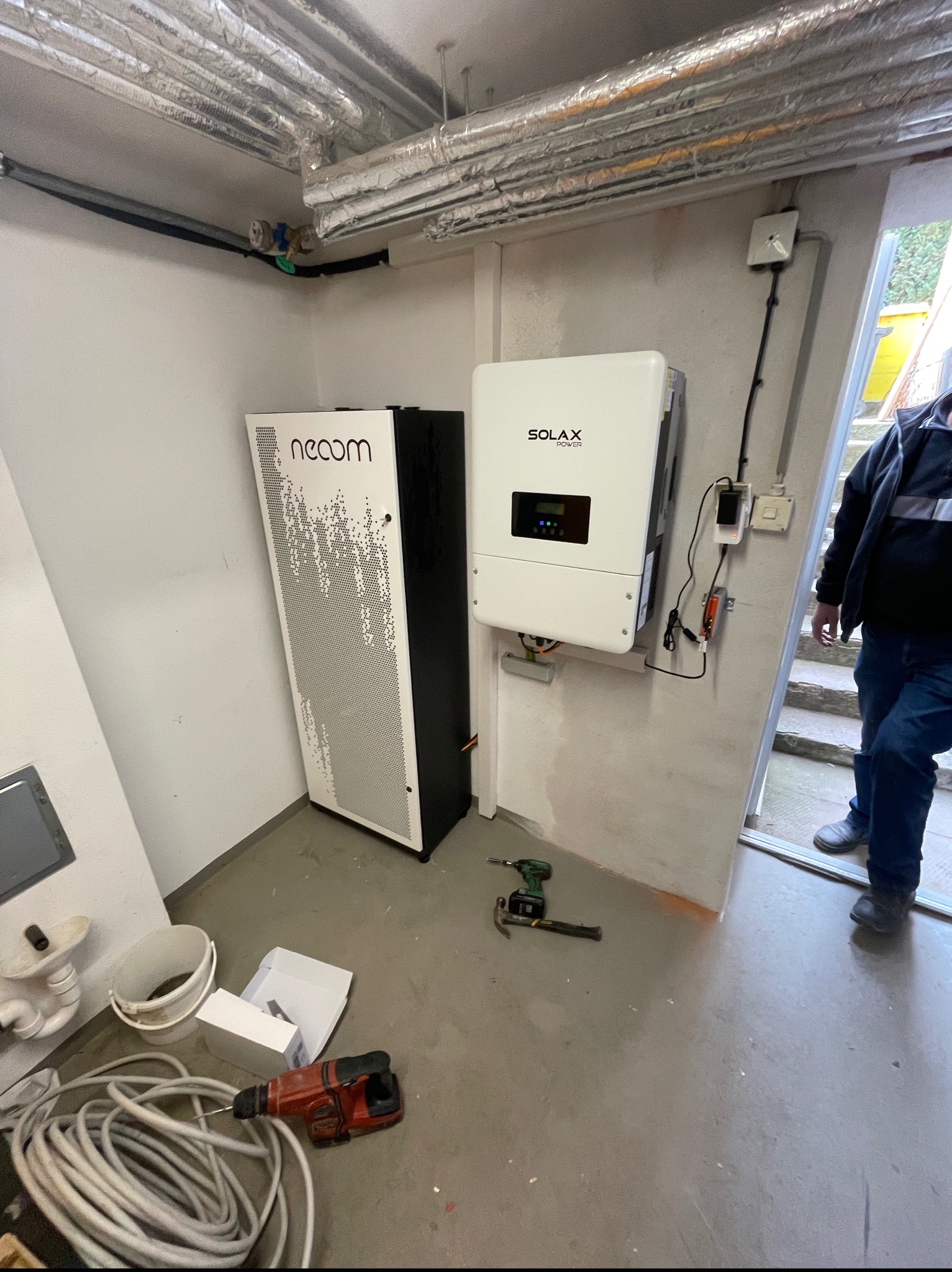 Wnętrze pomieszczenia technicznego z zainstalowanym magazynem energii Neoom i falownikiem Solax, widoczne narzędzia montażowe i fragment instalacji na suficie.