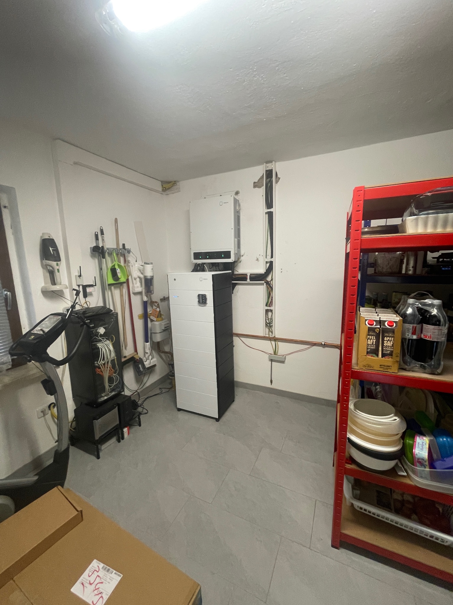 Biało-czarny moduł magazynu energii w garażu lub pomieszczeniu gospodarczym, widoczne okablowanie i osprzęt, obok regał z różnymi przedmiotami.