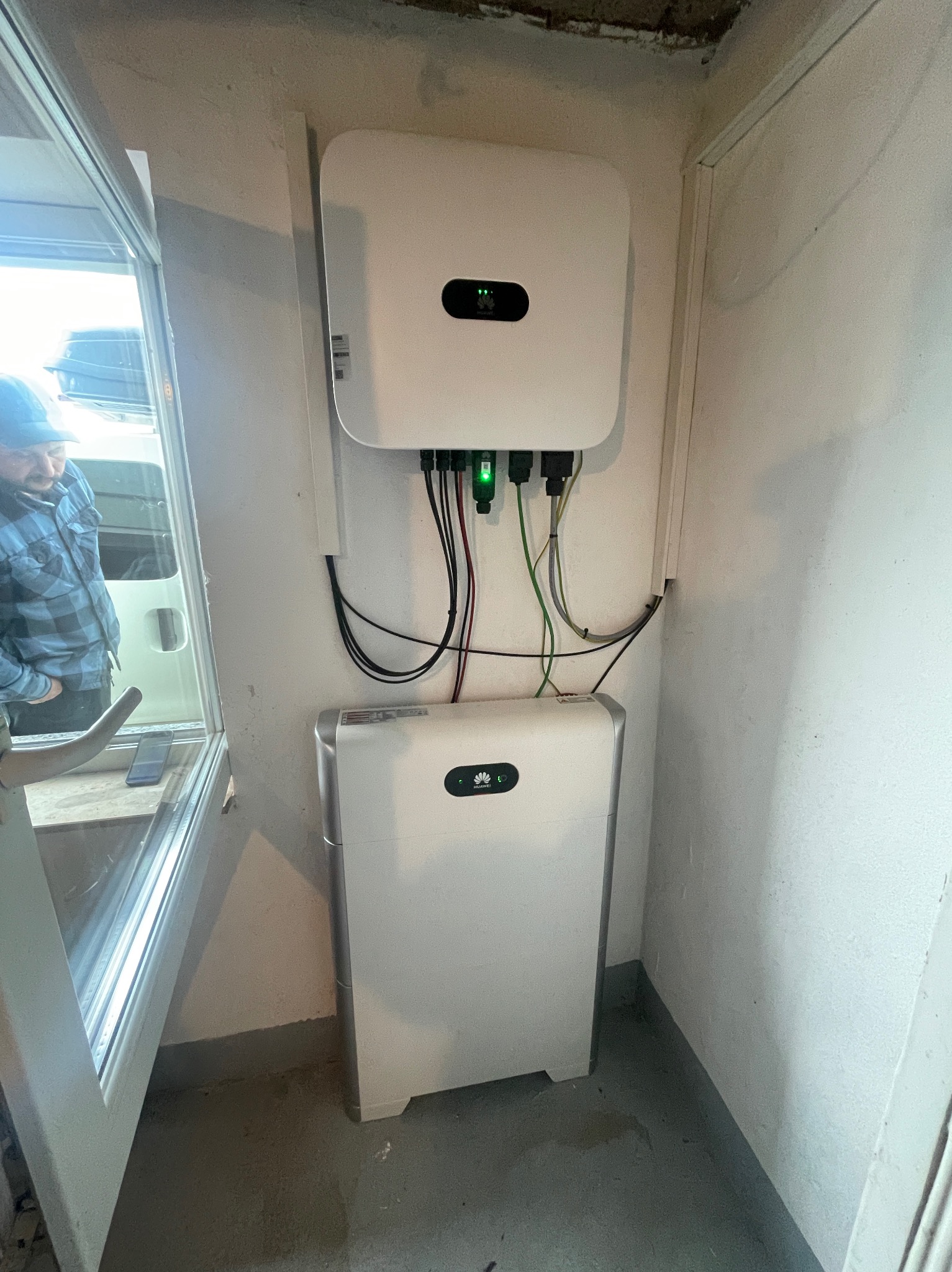Biały magazyn energii Huawei i inwerter zamontowane na ścianie w pomieszczeniu, widoczne kable połączeniowe, obok okno z widocznym fragmentem sylwetki osoby w niebieskiej koszuli w kratę.