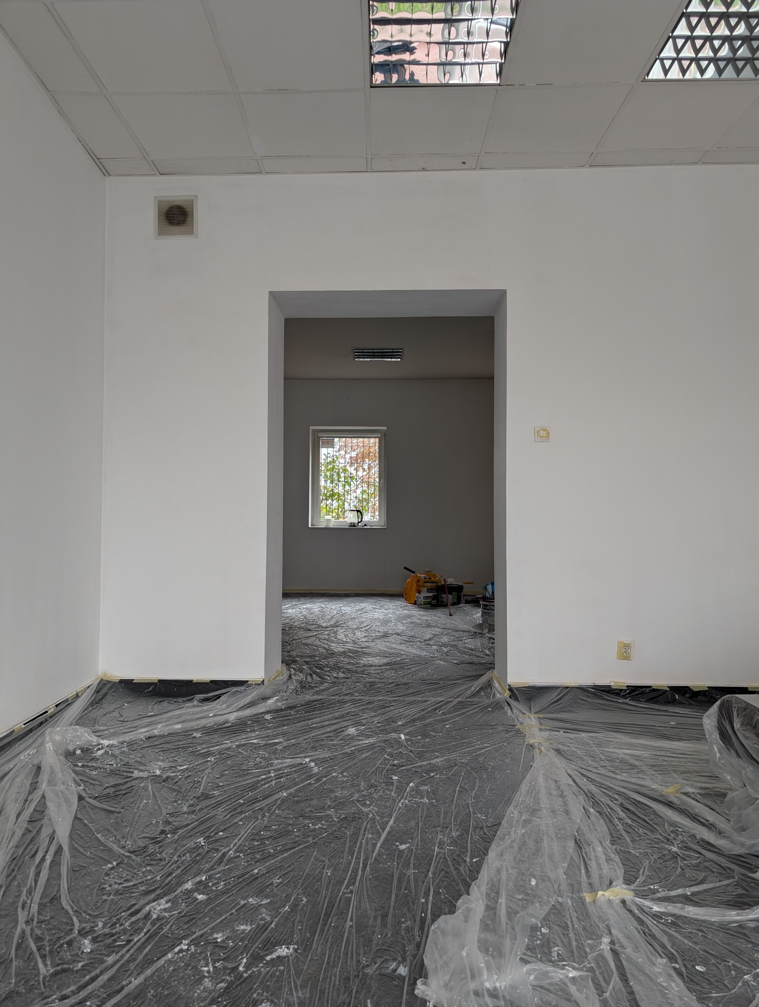 Przygotowane pomieszczenie biurowe do malowania: podłoga zabezpieczona folią, widoczne puszki z farbą i narzędzia malarskie w głębi pokoju, ściany świeżo pomalowane na biało, widok na okno...