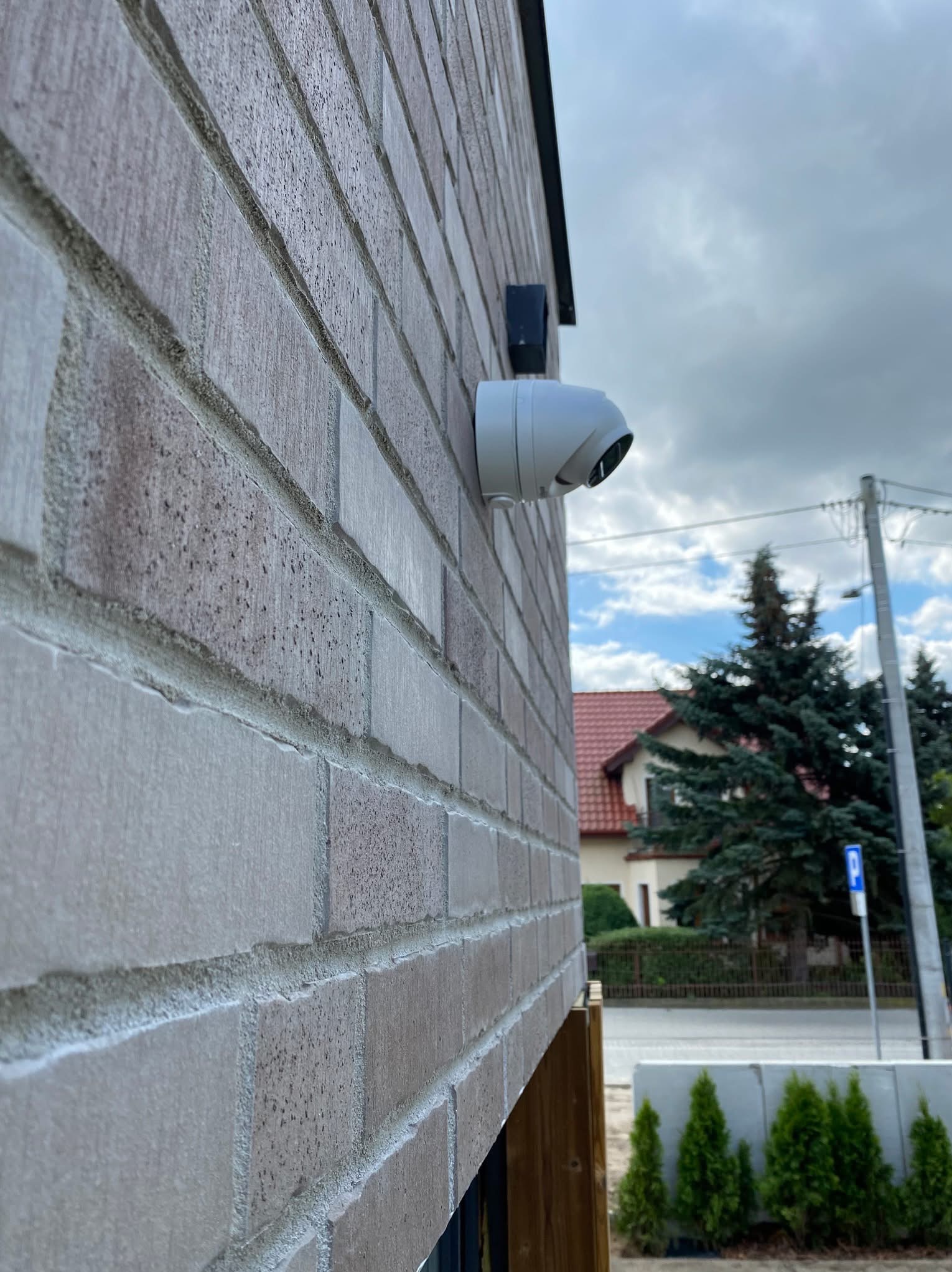 Kamera zewnętrzna, biała, kopułkowa, zamontowana na ceglanej ścianie budynku, widok z dołu, na tle pochmurnego nieba i fragmentu zabudowy.