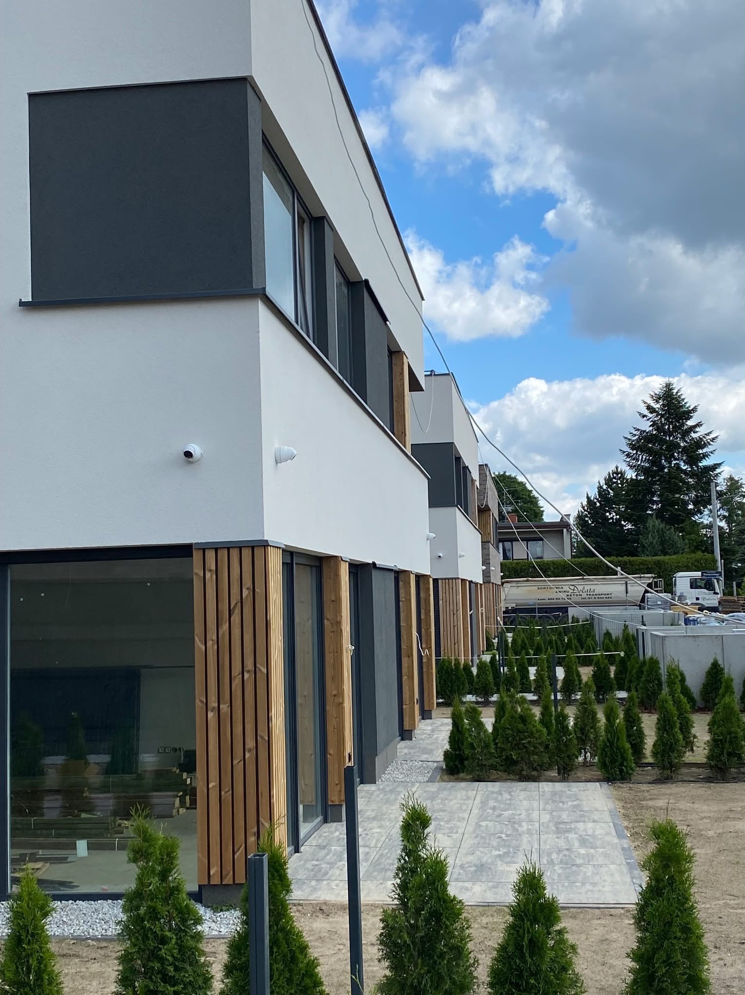Nowoczesny budynek z zainstalowaną kamerą monitoringu na elewacji, widoczne okna z drewnianymi panelami, małe drzewka iglaste i betonowa ścieżka.