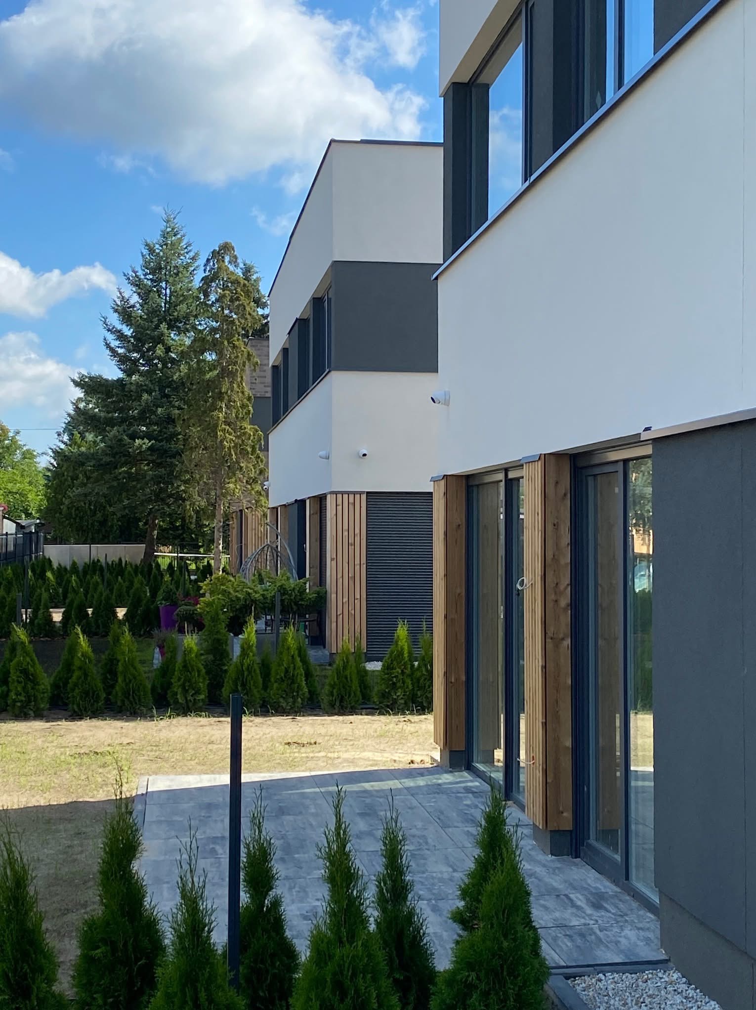 Nowoczesny dom z widocznymi kamerami monitoringu na elewacji, otoczony niskimi tujami i brukowanym chodnikiem, w tle wysokie drzewa i błękitne niebo z chmurami.