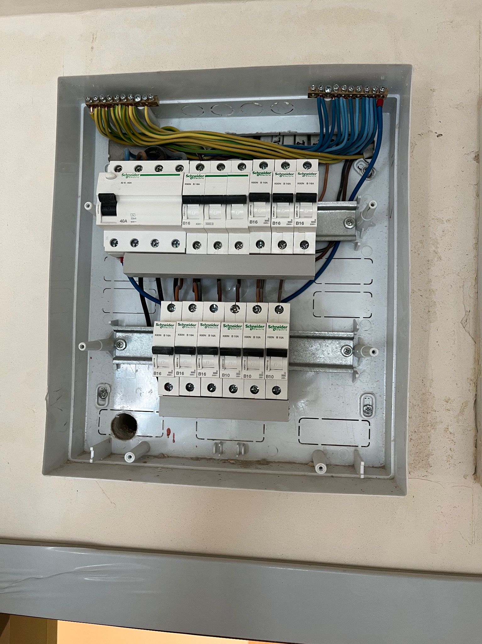 Otwarta skrzynka rozdzielcza z zamontowanymi bezpiecznikami Schneider Electric B16 i B10, widoczne podłączenia przewodów w kolorach niebieskim, żółto-zielonym i brązowym, zamocowane na szynach...
