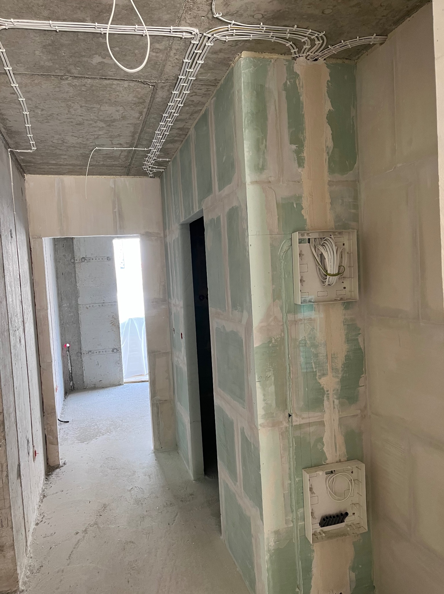 Wnętrze w stanie deweloperskim z widocznymi instalacjami elektrycznymi: kable prowadzone po suficie w białych listwach, puszki elektryczne na ścianach pokrytych zieloną płytą gipsowo-kartonową...