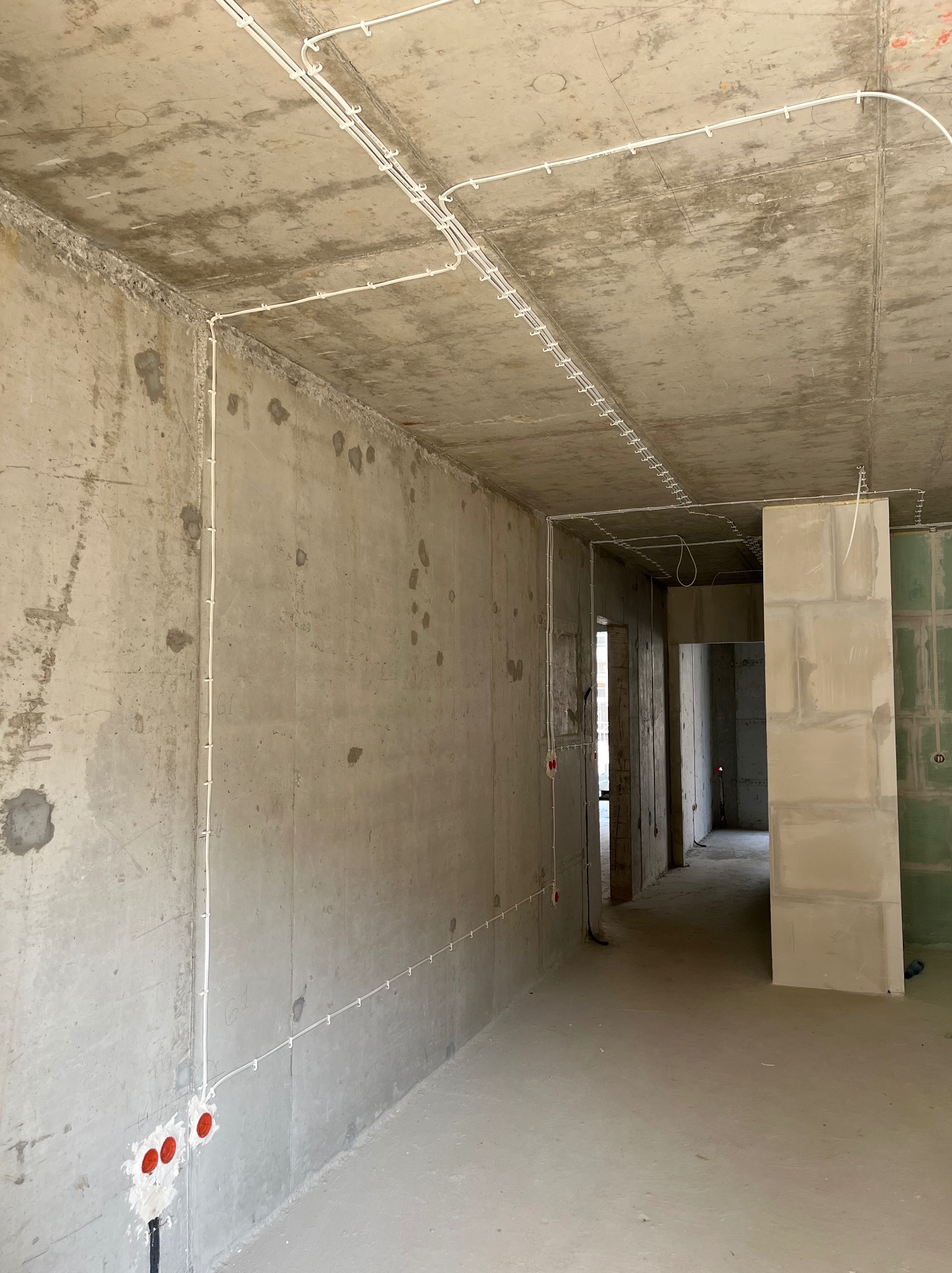 Surowy stan wnętrza z widocznymi przewodami elektrycznymi przytwierdzonymi do sufitu i ścian, gniazdka w puszkach podtynkowych w ścianie, betonowe ściany i podłoga, widok na kolejne pomieszczenia...
