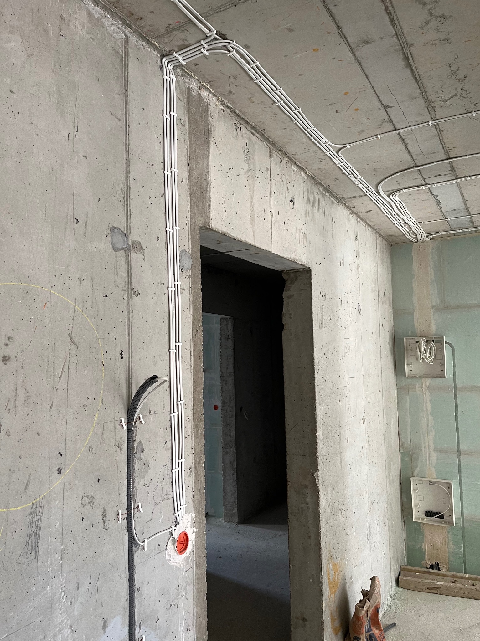 Surowy betonowy pokój w trakcie instalacji elektrycznej: trzy białe przewody prowadzone po ścianie i suficie, zabezpieczone uchwytami, widoczna puszka instalacyjna i fragment czarnego peszla.