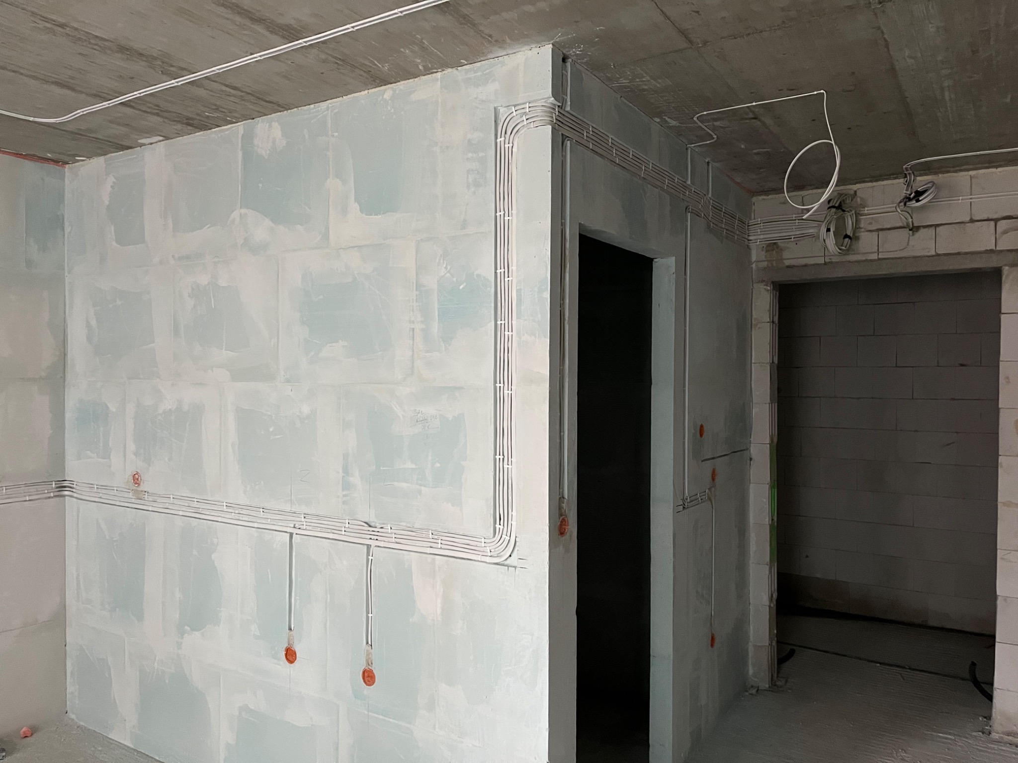 Instalacja elektryczna w surowym wnętrzu: białe przewody prowadzone po ścianach i suficie, widoczne puszki. Ściany wstępnie przygotowane do dalszych prac wykończeniowych.