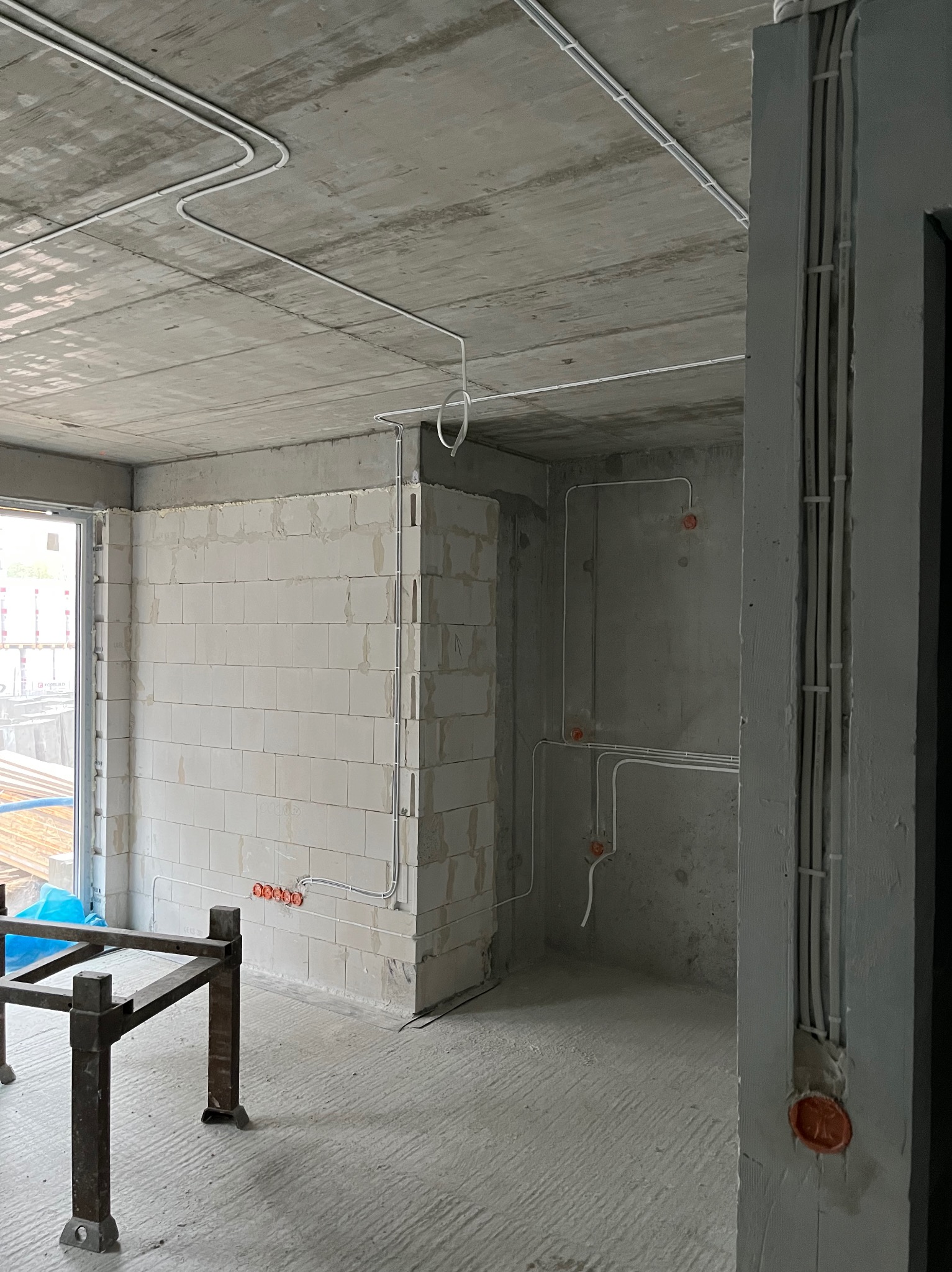 Surowy stan: widok na wnętrze z instalacją elektryczną w peszlach na ścianach i suficie. Pomarańczowe puszki elektryczne. Stan deweloperski z oknem.