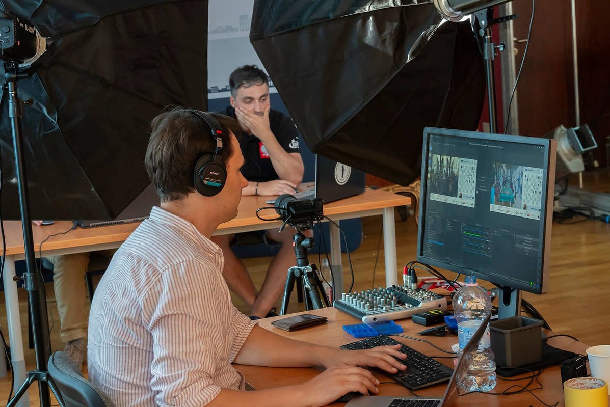 Mężczyzna w słuchawkach edytuje wideo na komputerze; widoczny mikser audio, oświetlenie studyjne i drugi mężczyzna w tle.