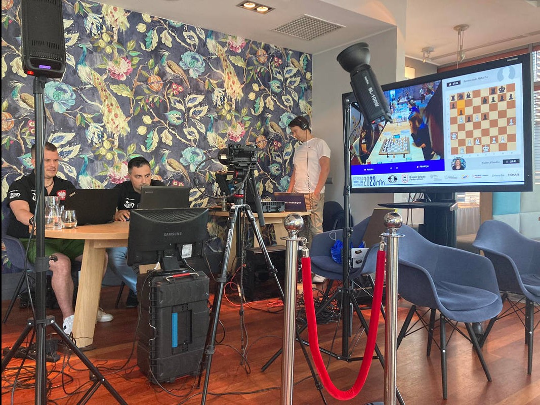 Ekipa filmowa podczas transmisji online turnieju szachowego, kamery na statywach, oświetlenie studyjne, monitory z podglądem rozgrywki, osoby pracujące przy laptopach, dekoracyjna tapeta w tle.