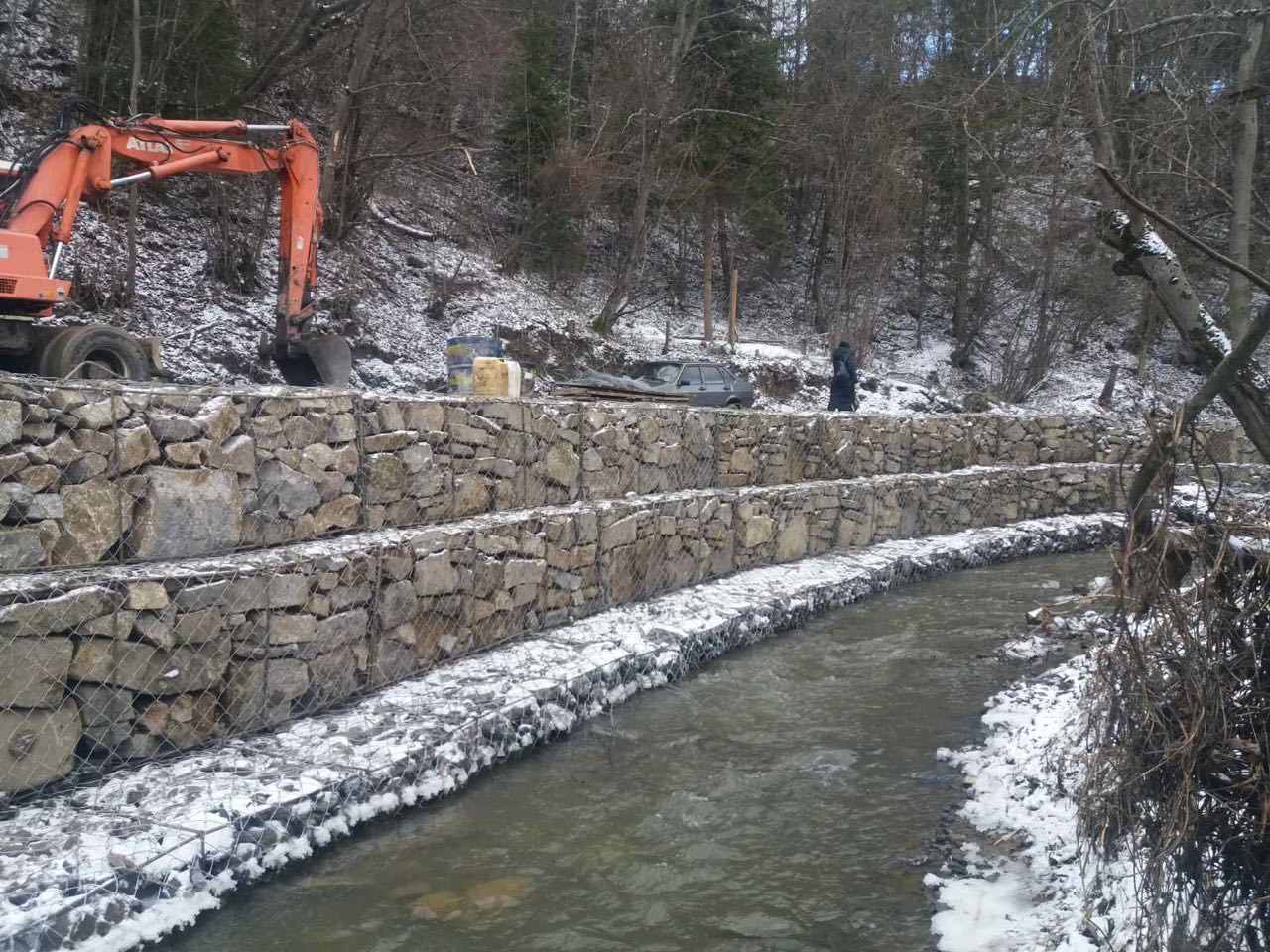 Wzmocnienie brzegów rzeki w Warszawie za pomocą gabionów, z widocznym pomarańczowym koparką i pracownikiem na tle zalesionego terenu.