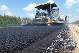 Rozkładanie świeżego asfaltu przez maszynę na budowie drogi, widoczne ślady opon i równa powierzchnia, w tle drzewa i błękitne niebo z chmurami.