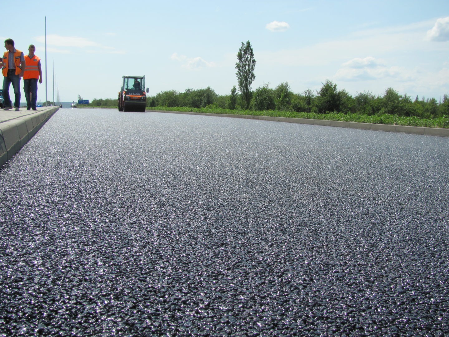 Świeżo wylany asfalt na drodze, walec drogowy w oddali, dwóch pracowników w kamizelkach odblaskowych przy krawężniku.