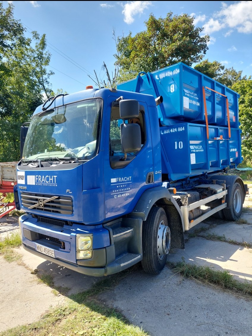 Niebieska ciężarówka z dwoma kontenerami na platformie, z widocznym logo firmy „FRACHT” i numerami kontaktowymi, zaparkowana na betonowym placu w słoneczny dzień.