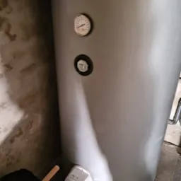 Szary zasobnik ciepłej wody użytkowej z widocznym termometrem i elementami instalacji, stojący przy szarej, surowej ścianie.