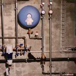 HYDRO-INSTAL-TECH SPÓŁKA Z OGRANICZONĄ ODPOWIEDZIALNOŚCIĄ - Kompleksowa instalacja grzewcza z rur stalowych, manometrami i niebieskim zbiornikiem wyrównawczym na tle surowej, betonowej ściany.