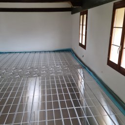 HYDRO-INSTAL-TECH SPÓŁKA Z OGRANICZONĄ ODPOWIEDZIALNOŚCIĄ - Przygotowanie do instalacji ogrzewania podłogowego: metalowa siatka rozłożona na folii izolacyjnej w przestronnym pomieszczeniu z drewnianymi oknami i belkami stropowymi.