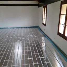 Przygotowanie do instalacji ogrzewania podłogowego: metalowa siatka rozłożona na folii izolacyjnej w przestronnym pomieszczeniu z drewnianymi oknami i belkami stropowymi.