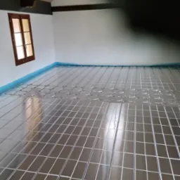 Montaż ogrzewania podłogowego: widok na rozłożoną siatkę metalową z rurami w pomieszczeniu z oknem i drewnianymi belkami stropowymi, przygotowaną do zalania wylewką.