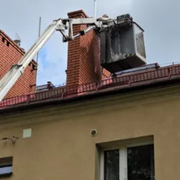 Podnośnik koszowy przy kominie na dachu budynku. Prace na wysokościach, widoczny fragment elewacji i okno. Szaro-białe niebo w tle.