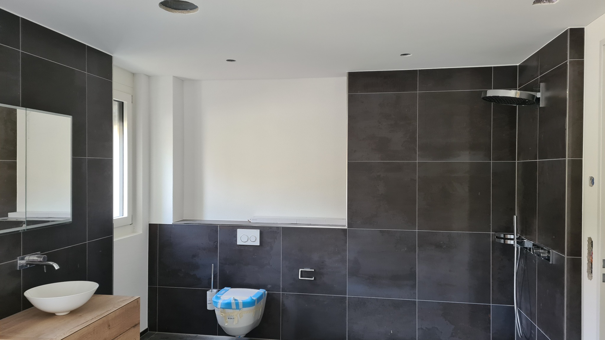 Nowoczesna łazienka w trakcie wykończenia, z ciemnymi płytkami, białą umywalką na drewnianej szafce, zabezpieczoną toaletą i chromowaną armaturą prysznicową.