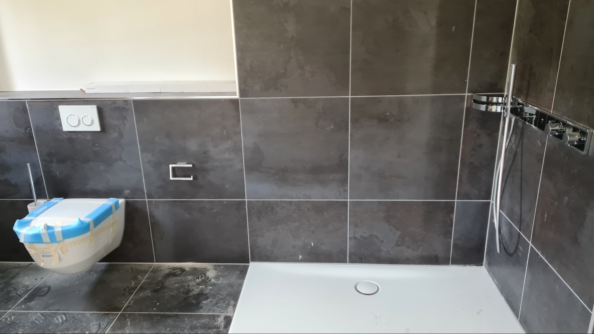 Nowoczesna łazienka w trakcie remontu: szara, kwadratowa glazura, biały brodzik i podwieszana toaleta zabezpieczona taśmą malarską, widoczne elementy instalacji prysznicowej.