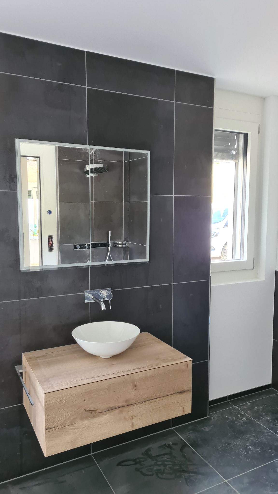 Nowoczesna łazienka w Ustroniu z czarnymi płytkami, umywalką nablatową na drewnianej szafce i lustrem w zabudowie, odzwierciedlającym kabinę prysznicową.