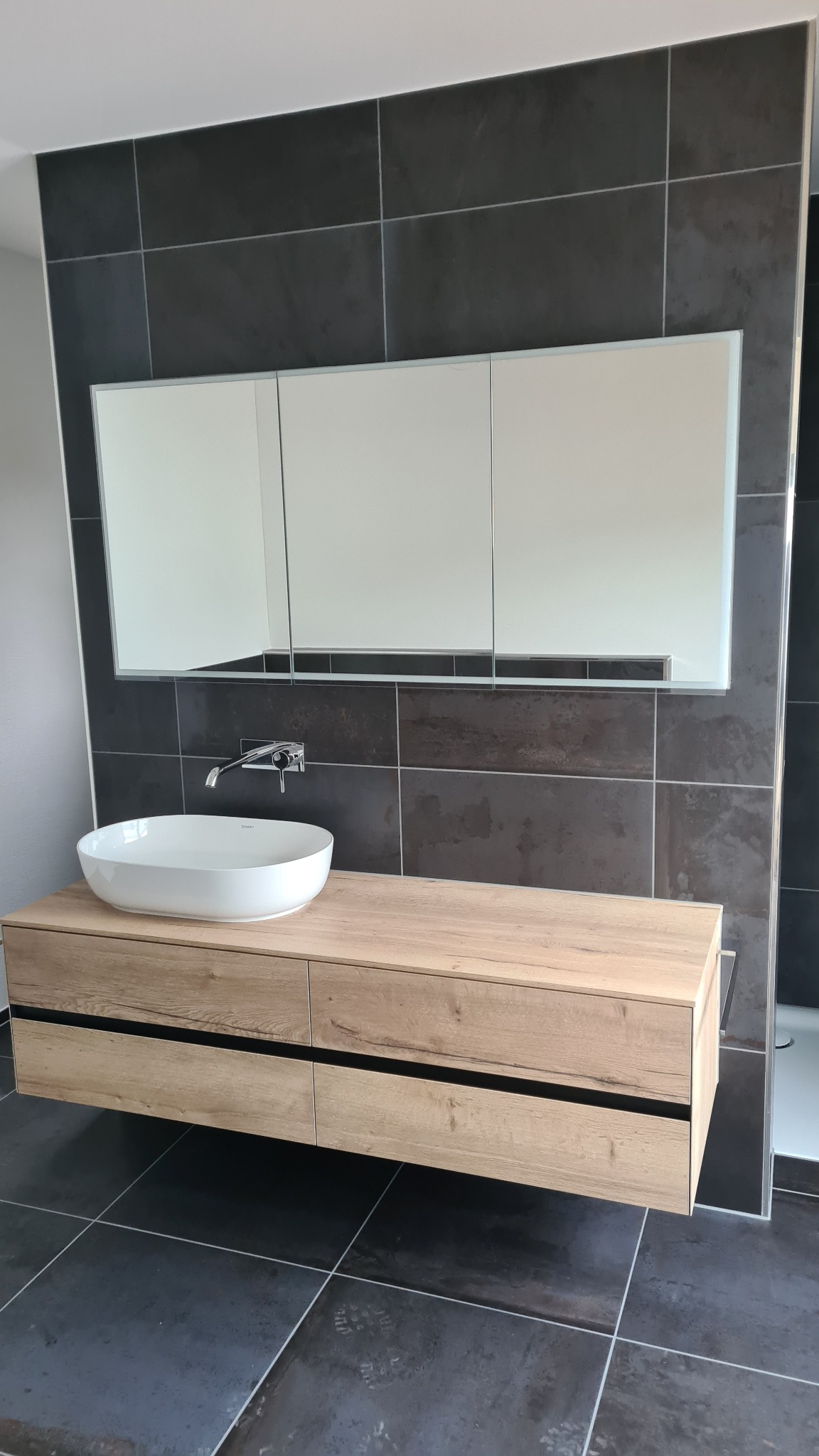 Nowoczesna łazienka z wiszącą szafką z umywalką nablatową, lustrem z szafkami i ciemnymi płytkami na ścianach i podłodze.