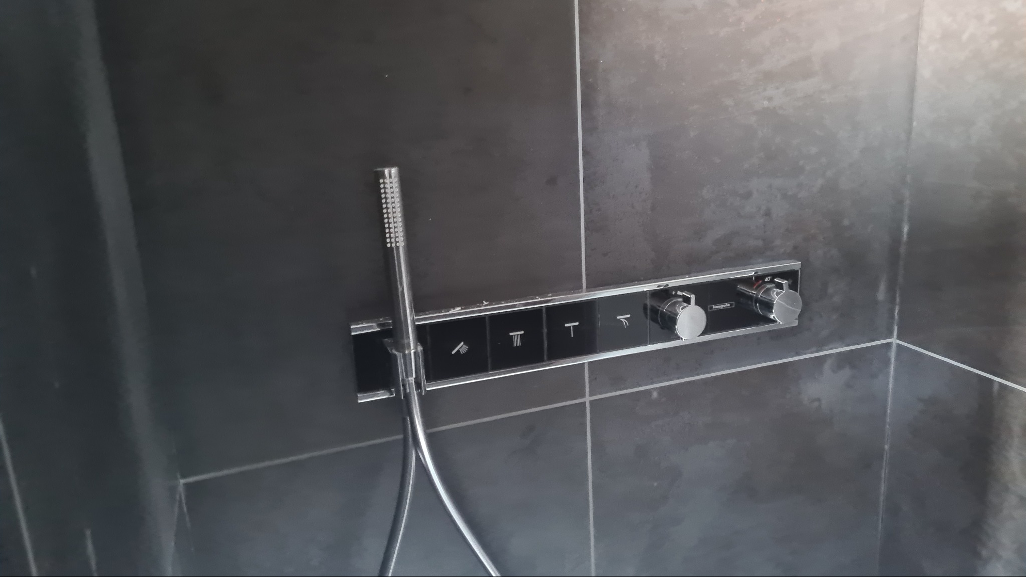 Nowoczesna bateria prysznicowa z panelem sterowania na ciemnych, matowych płytkach, widoczna słuchawka prysznicowa z chromowanym wykończeniem i oznaczenia funkcji na panelu.