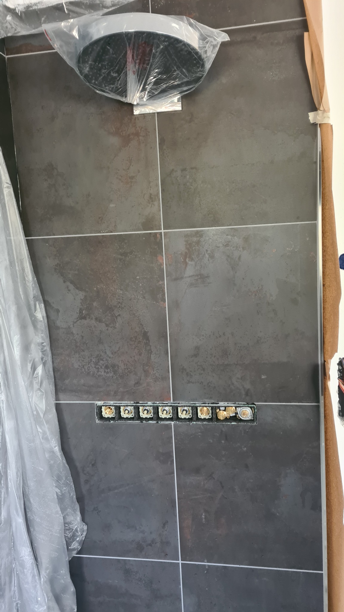 Szare płytki ścienne z widocznymi otworami na przyłącza instalacji hydraulicznej, częściowo zakryte folią ochronną i zabezpieczeniami budowlanymi.