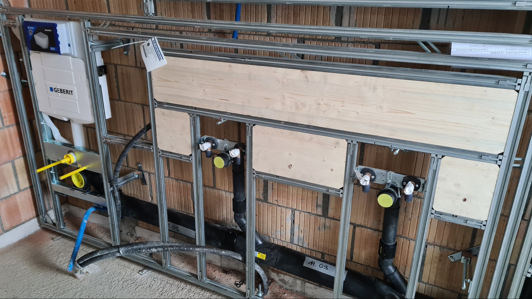 Stelaż instalacyjny podtynkowy z elementami instalacji wodno-kanalizacyjnej przed zabudową, widoczne rury, zawory, elementy mocujące i płyty drewniane.