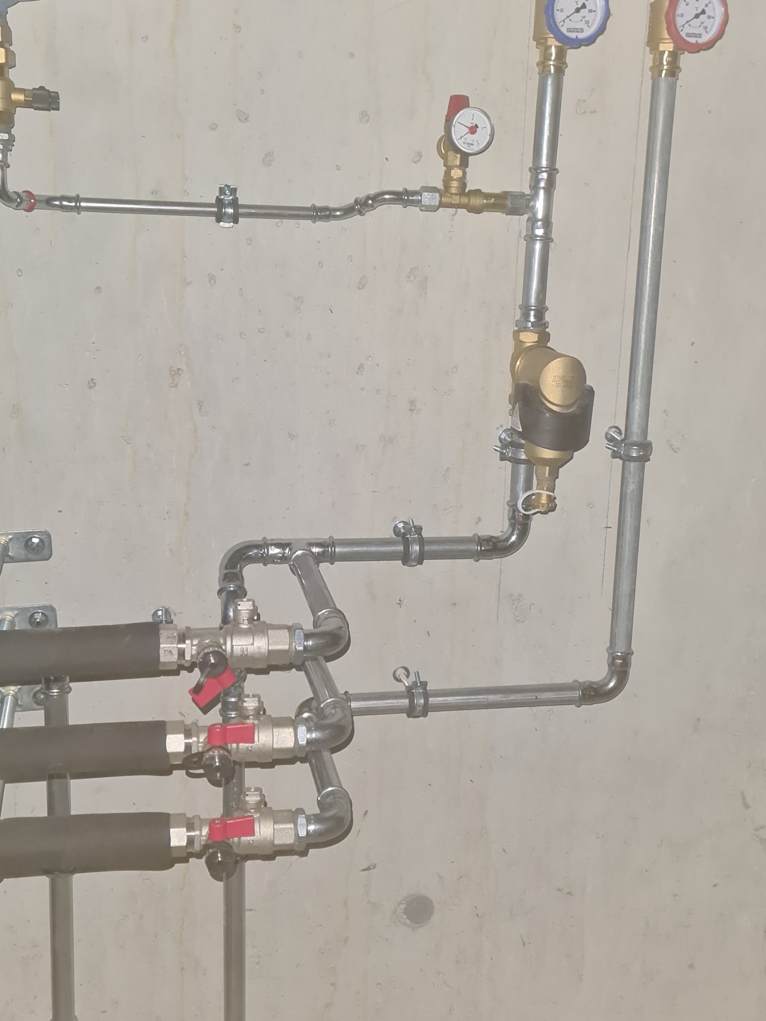 Szczegółowe ujęcie instalacji hydraulicznej z rurami, zaworami z czerwonymi pokrętłami, manometrami i izolacją termiczną na tle surowej ściany betonowej.