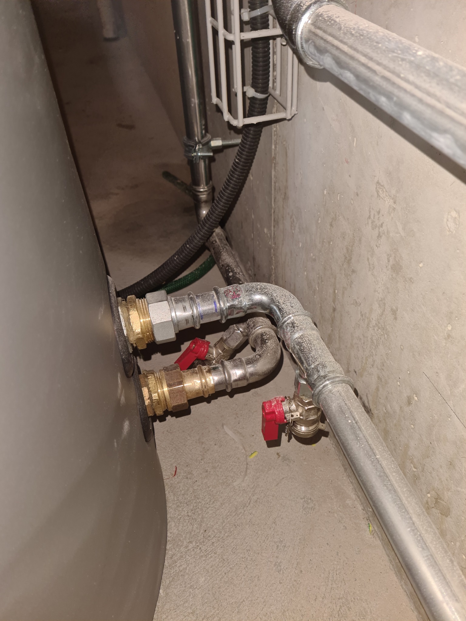 Szczegółowe ujęcie podłączenia rur z zaworami z czerwonymi uchwytami do zbiornika, prawdopodobnie bojlera, na tle surowej ściany i podłogi w piwnicy.