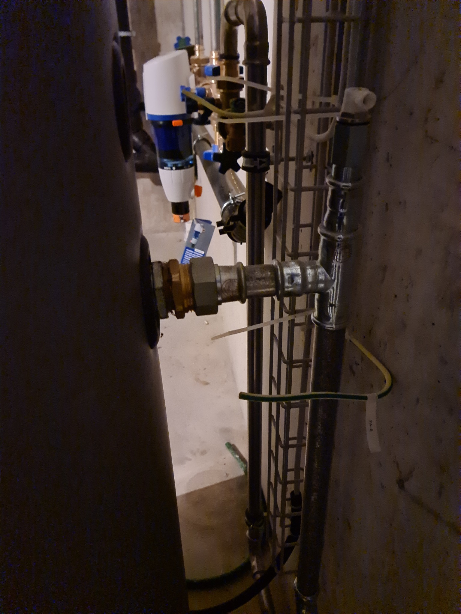 Fragment kotłowni z widocznym kotłem, filtrem wody, rurami instalacji grzewczej zabezpieczonymi siatką i zaworami, w surowym pomieszczeniu z betonową ścianą.