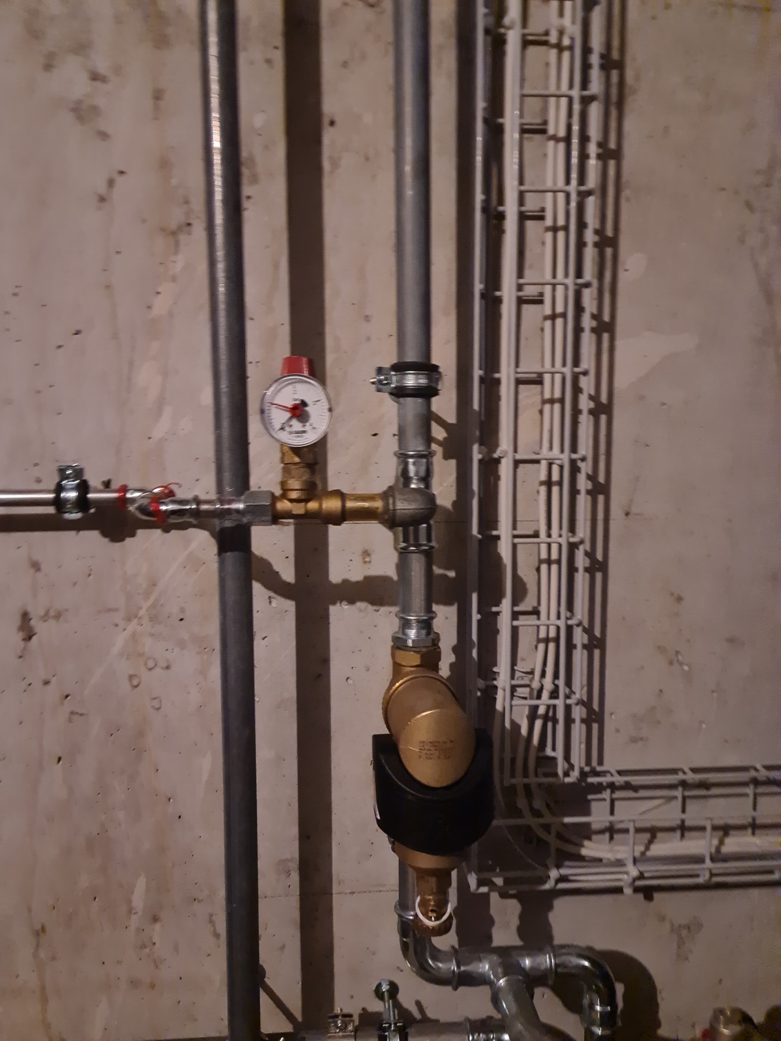 Fragment instalacji gazowej z manometrem, zaworami i filtrem, widoczne rury stalowe i miedziane na tle surowej ściany z korytkami kablowymi.