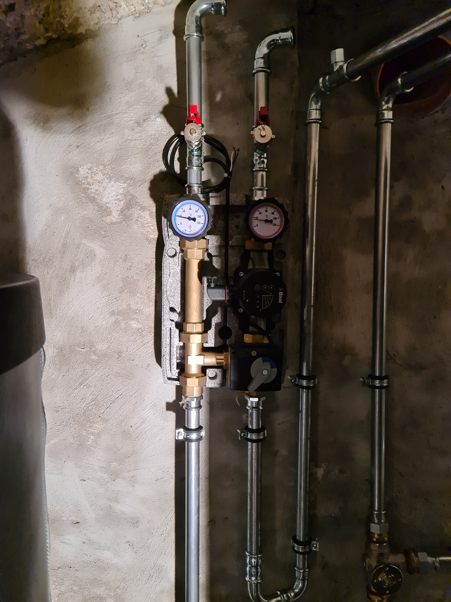 Dwie pionowe instalacje rurowe z manometrami i pompami obiegowymi na szorstkiej, betonowej ścianie. Widoczne złączki, zawory i mocowania rur.