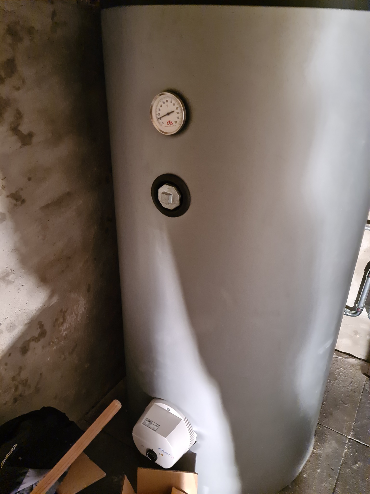 Szary zasobnik ciepłej wody użytkowej z widocznym termometrem i elementami instalacji, stojący przy szarej, surowej ścianie.