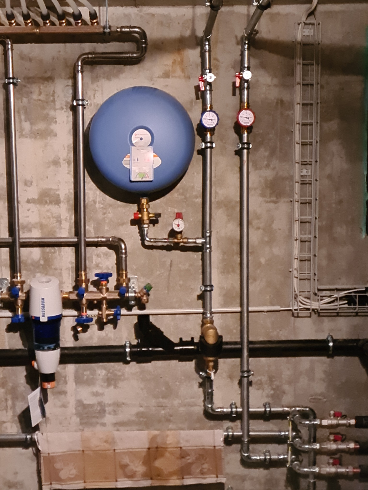 Kompleksowa instalacja grzewcza z rur stalowych, manometrami i niebieskim zbiornikiem wyrównawczym na tle surowej, betonowej ściany.